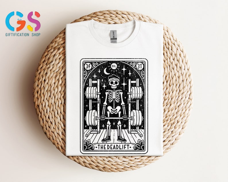 May include: White t-shirt featuring a black and white illustration of a skeleton lifting weights. The illustration is framed in a black border with the text "The Deadlift" at the bottom.