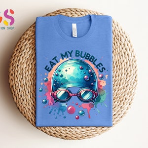 May include: A light blue t-shirt with a colorful graphic of a swimmer's cap and goggles with the text "Eat My Bubbles".