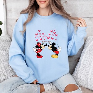 May include: Light blue sweatshirt featuring a graphic of Mickey and Minnie Mouse kissing, with red hearts floating above them. Minnie is wearing a red polka dot dress and bow. Mickey is in red shorts and yellow shoes. The sweatshirt is a comfortable, casual style.