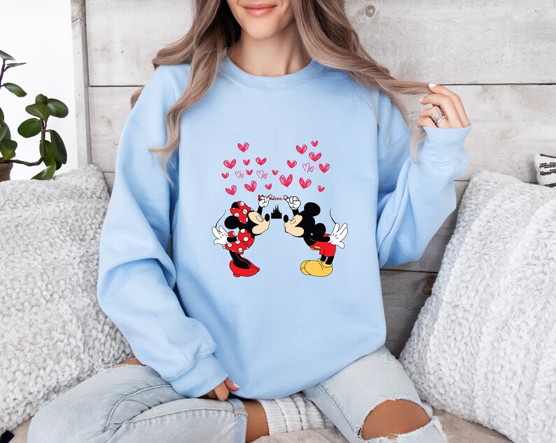 May include: Light blue sweatshirt featuring a graphic of Mickey and Minnie Mouse kissing, with red hearts floating above them. Minnie is wearing a red polka dot dress and bow. Mickey is in red shorts and yellow shoes. The sweatshirt is a comfortable, casual style.