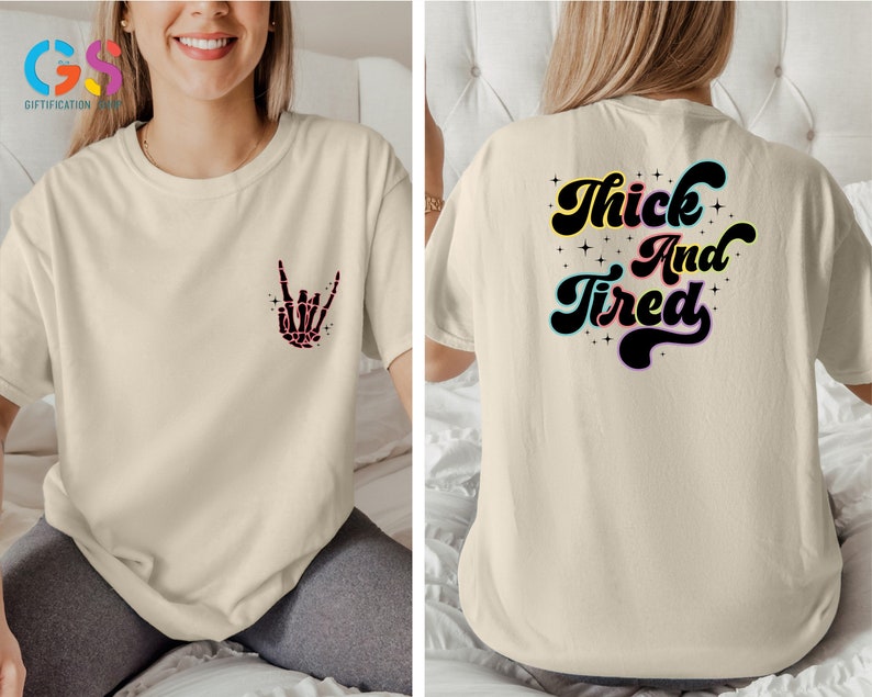 Op de afbeelding: Een beige T-shirt met een skelethand die een rock-and-roll-teken maakt op de voorkant en de tekst "Thick And Tired" in een retro-lettertype op de achterkant.