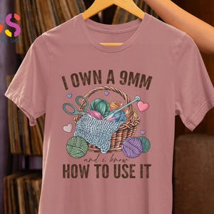 May include: A pink t-shirt with the text "I own a 9mm and I know how to use it". The text is in brown and there is an illustration of a basket of yarn with scissors and needles.