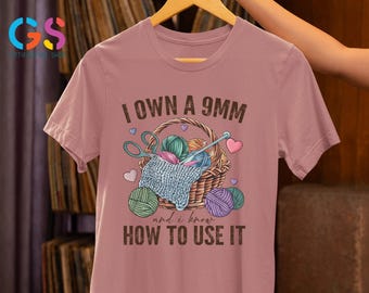 May include: A pink t-shirt with the text "I own a 9mm and I know how to use it". The text is in brown and there is an illustration of a basket of yarn with scissors and needles.