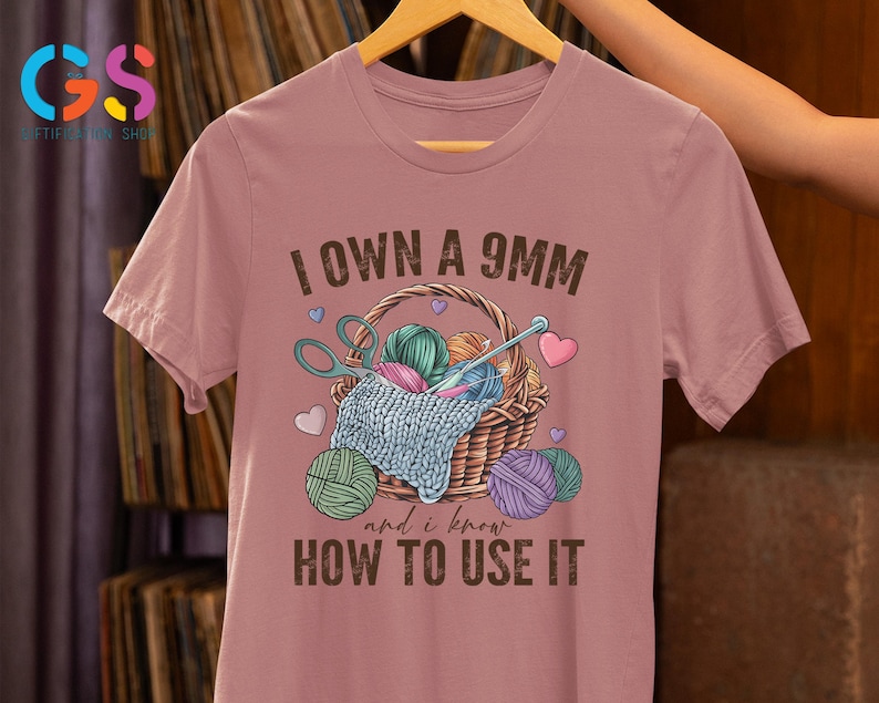 May include: A pink t-shirt with the text "I own a 9mm and I know how to use it". The text is in brown and there is an illustration of a basket of yarn with scissors and needles.