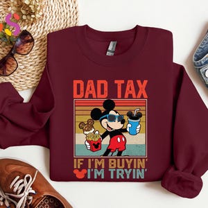 May include: A maroon sweatshirt featuring a graphic of Mickey Mouse wearing sunglasses and holding a drink and fries. The text "DAD TAX" and "IF I'M BUYIN' I'M TRYIN'" are printed above and below the graphic in a vintage style.