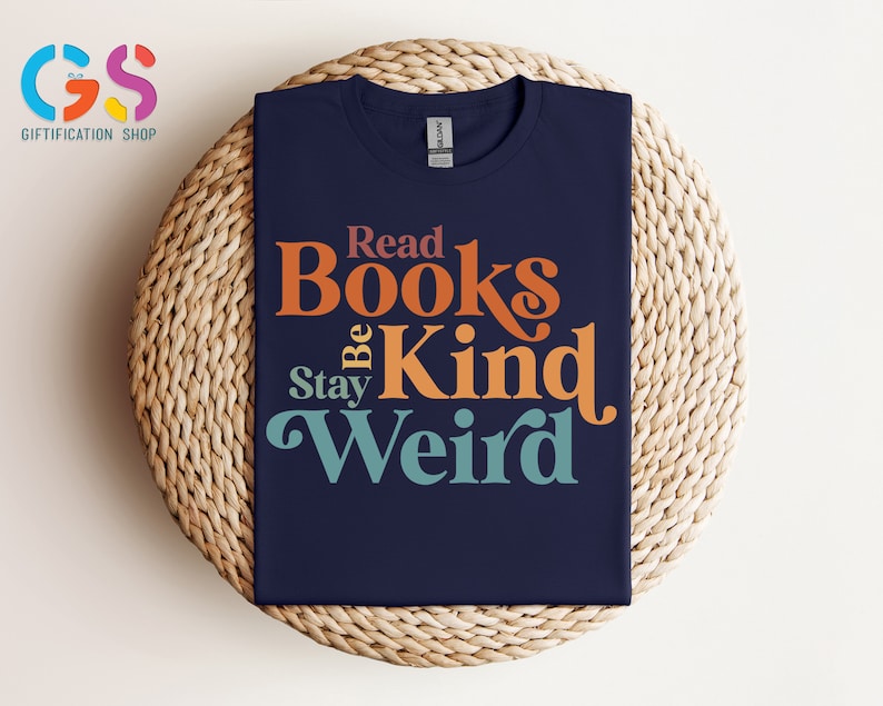 K&ouml;nnte beinhalten: Ein marineblaues T-Shirt mit einer Retro-Grafik, auf der "Read Books Be Kind Stay Weird" steht. Der Text ist in Orange, Gelb und Blau.