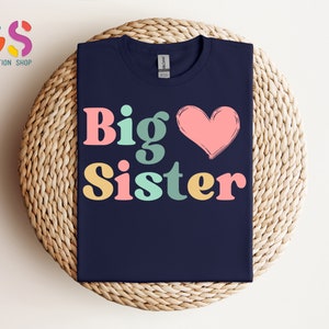 May include: A navy blue t-shirt with the text "Big Sister" in colorful retro-style letters and a pink heart.