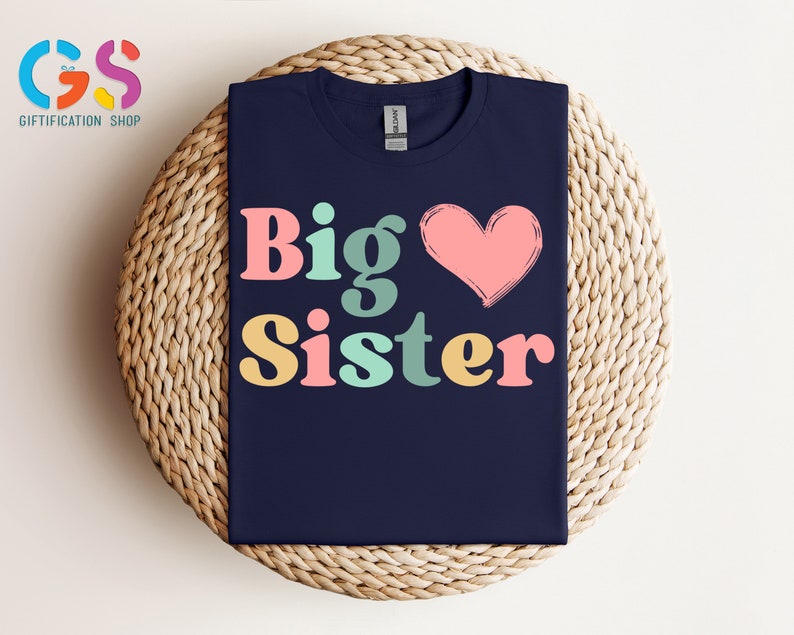 May include: A navy blue t-shirt with the text "Big Sister" in colorful retro-style letters and a pink heart.