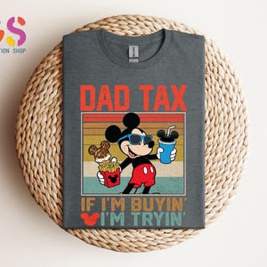 May include: Dark gray t-shirt featuring Mickey Mouse wearing sunglasses and holding a drink, fries, and ice cream. The shirt has the text "DAD TAX" and "IF I'M BUYIN' I'M TRYIN'". The design is in red, yellow, and blue.