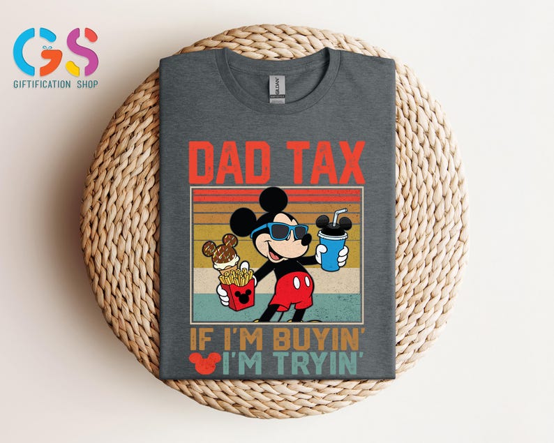 May include: Dark gray t-shirt featuring Mickey Mouse wearing sunglasses and holding a drink, fries, and ice cream. The shirt has the text "DAD TAX" and "IF I'M BUYIN' I'M TRYIN'". The design is in red, yellow, and blue.