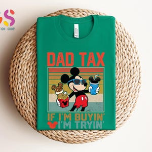 May include: Green t-shirt featuring a graphic of Mickey Mouse wearing sunglasses and holding food and a drink. The text "DAD TAX" is above the graphic, and "IF I'M BUYIN' I'M TRYIN'" is below. The shirt is displayed on a woven surface.