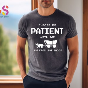 Puede incluir: Una camiseta gris oscuro con un dise&ntilde;o gr&aacute;fico blanco. El gr&aacute;fico presenta una imagen pixelada de un carro cubierto tirado por un caballo. El texto en la camiseta dice "Please be patient with me I'm from the 1900s".