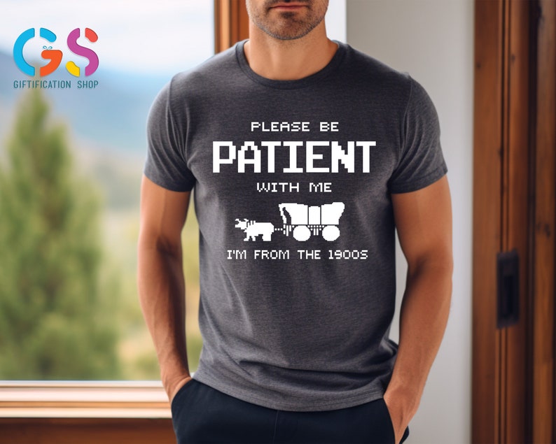 Puede incluir: Una camiseta gris oscuro con un dise&ntilde;o gr&aacute;fico blanco. El gr&aacute;fico presenta una imagen pixelada de un carro cubierto tirado por un caballo. El texto en la camiseta dice "Please be patient with me I'm from the 1900s".