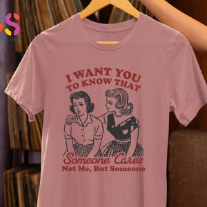 Op de afbeelding: Een roze T-shirt met een vintage-stijl grafische afbeelding van twee vrouwen. De tekst op het shirt luidt "Ik wil dat je weet dat iemand geeft, niet ik, maar iemand."