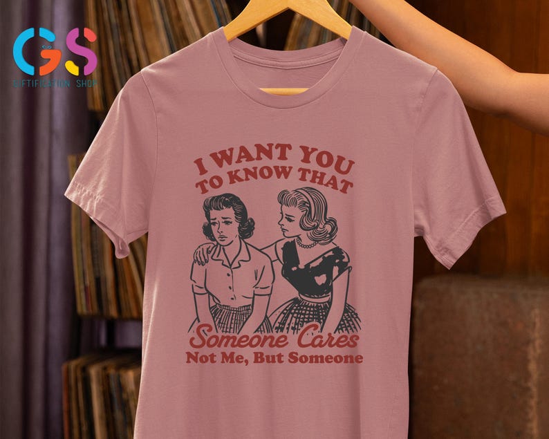 Op de afbeelding: Een roze T-shirt met een vintage-stijl grafische afbeelding van twee vrouwen. De tekst op het shirt luidt "Ik wil dat je weet dat iemand geeft, niet ik, maar iemand."