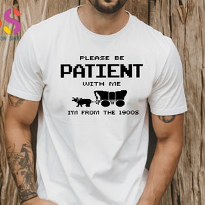 Puede incluir: Camiseta blanca con texto negro que dice "Por favor, ten paciencia conmigo, soy de los a&ntilde;os 1900". El texto est&aacute; en una fuente pixelada. Debajo del texto hay una ilustraci&oacute;n en blanco y negro de un carro tirado por un toro.