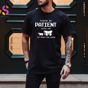 Puede incluir: Camiseta negra con texto blanco que dice "Por favor, ten paciencia conmigo, soy de los a&ntilde;os 1900". El texto est&aacute; en una fuente pixelada. Un peque&ntilde;o gr&aacute;fico de un carro tirado por caballos est&aacute; debajo del texto.