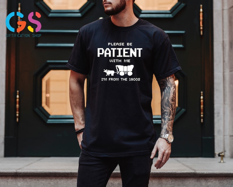 Puede incluir: Camiseta negra con texto blanco que dice "Por favor, ten paciencia conmigo, soy de los a&ntilde;os 1900". El texto est&aacute; en una fuente pixelada. Un peque&ntilde;o gr&aacute;fico de un carro tirado por caballos est&aacute; debajo del texto.