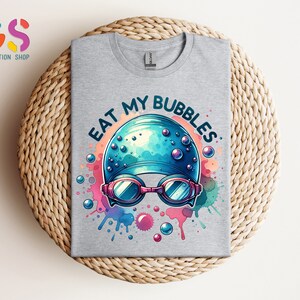 May include: A gray t-shirt with a blue and white swimming cap and goggles with the text "Eat My Bubbles" in a colorful watercolor design.