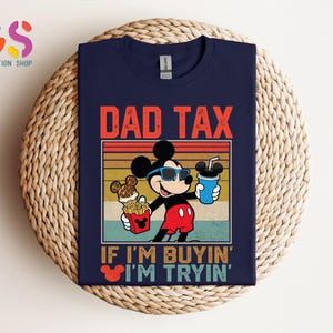 May include: Navy blue t-shirt featuring a graphic of Mickey Mouse wearing sunglasses and holding food and a drink. The text "DAD TAX" is above the graphic, with "IF I'M BUYIN' I'M TRYIN'" below. The shirt is displayed on a woven surface.