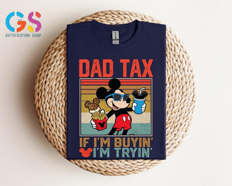 May include: Navy blue t-shirt featuring a graphic of Mickey Mouse wearing sunglasses and holding food and a drink. The text "DAD TAX" is above the graphic, with "IF I'M BUYIN' I'M TRYIN'" below. The shirt is displayed on a woven surface.