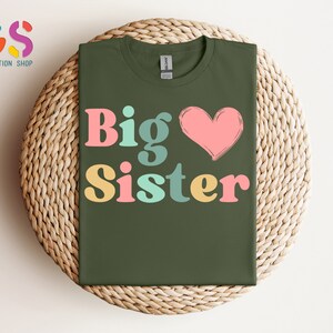 May include: A green t-shirt with the text "Big Sister" in colorful letters and a pink heart.