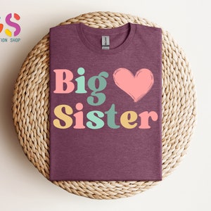 May include: A maroon t-shirt with the text "Big Sister" in colorful retro-style letters and a pink heart.