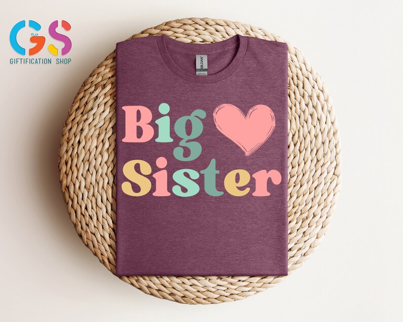 May include: A maroon t-shirt with the text "Big Sister" in colorful retro-style letters and a pink heart.