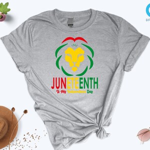 Pode incluir: Uma camiseta cinza mesclada com um design gr&aacute;fico colorido. O design apresenta uma cabe&ccedil;a de le&atilde;o amarela, cercada por formas verdes e vermelhas, com as palavras "JUNETEENTH" e "Is My Freedom Day" em v&aacute;rias cores.