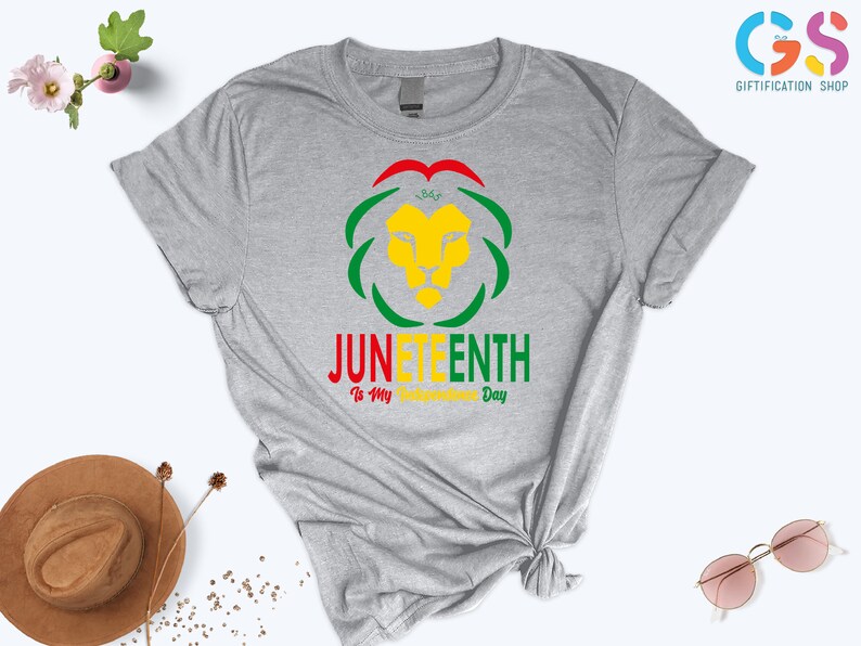 Pode incluir: Uma camiseta cinza mesclada com um design gr&aacute;fico colorido. O design apresenta uma cabe&ccedil;a de le&atilde;o amarela, cercada por formas verdes e vermelhas, com as palavras "JUNETEENTH" e "Is My Freedom Day" em v&aacute;rias cores.