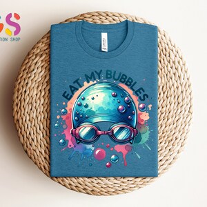 May include: A blue t-shirt with a watercolor design of a swimmer's cap and goggles. The text "Eat My Bubbles" is written in a white outline around the design.