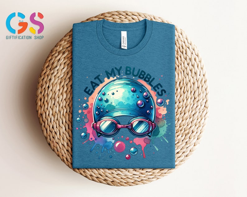 May include: A blue t-shirt with a watercolor design of a swimmer's cap and goggles. The text "Eat My Bubbles" is written in a white outline around the design.