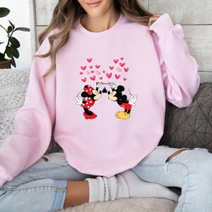 May include: A light pink sweatshirt featuring a graphic of Mickey and Minnie Mouse kissing, with red heart accents. The characters are in their classic outfits. The word "Believe" is written above the castle. A cozy, casual top.