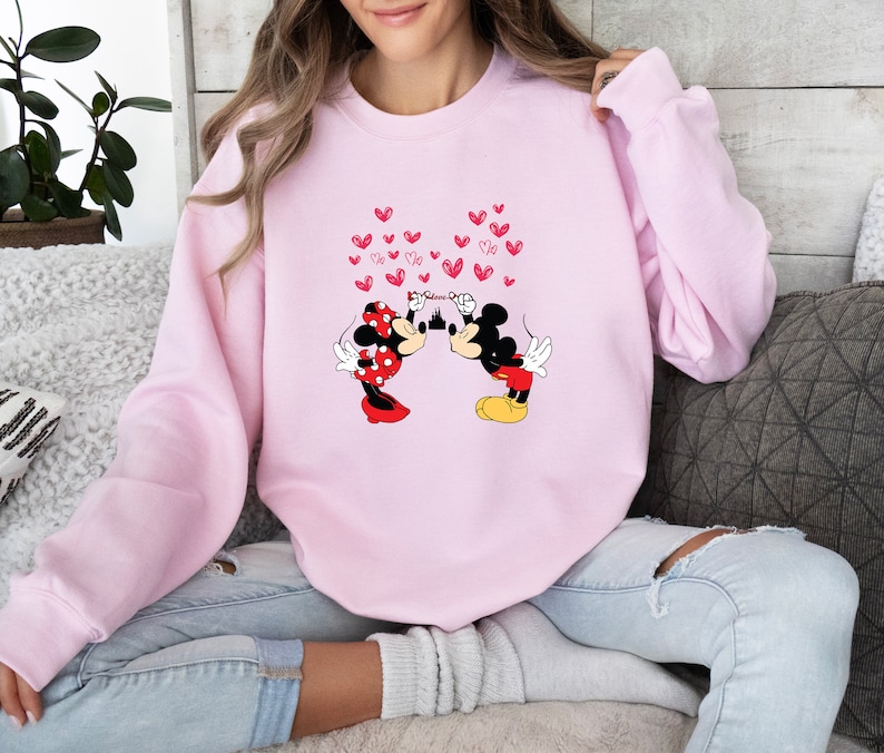 May include: A light pink sweatshirt featuring a graphic of Mickey and Minnie Mouse kissing, with red heart accents. The characters are in their classic outfits. The word "Believe" is written above the castle. A cozy, casual top.