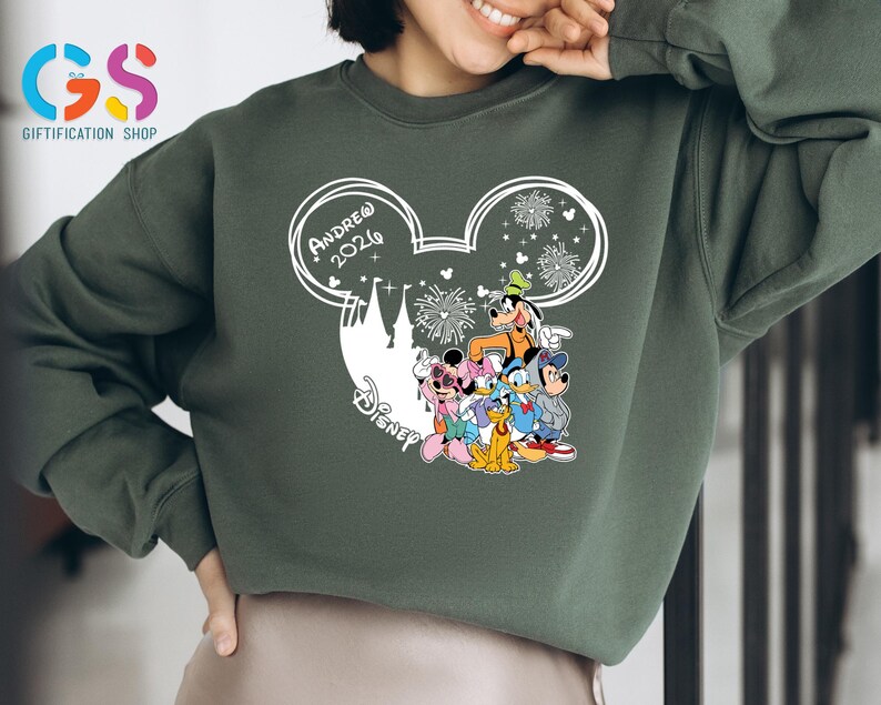 Pode incluir: Camisola verde com um design branco da cabe&ccedil;a do Mickey Mouse, o texto "Andrew 2026" e personagens da Disney. O design inclui fogos de artif&iacute;cio e uma silhueta de castelo. A camisola &eacute; de cor verde lisa.