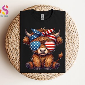 Puede incluir: Camiseta negra con un dibujo de una vaca de las Tierras Altas con gafas de sol y una diadema, ambas decoradas con la bandera americana. La vaca tiene pelaje marr&oacute;n y un aro en la nariz. La camiseta se muestra sobre una superficie tejida.