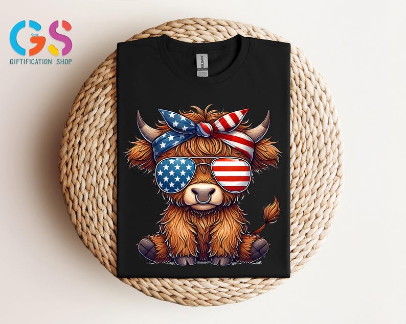 Puede incluir: Camiseta negra con un dibujo de una vaca de las Tierras Altas con gafas de sol y una diadema, ambas decoradas con la bandera americana. La vaca tiene pelaje marr&oacute;n y un aro en la nariz. La camiseta se muestra sobre una superficie tejida.