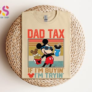 May include: Beige t-shirt featuring a graphic of Mickey Mouse wearing sunglasses and holding a drink and fries. The text "DAD TAX" is above the graphic, and "IF I'M BUYIN' I'M TRYIN'" is below. The shirt is on a woven surface.
