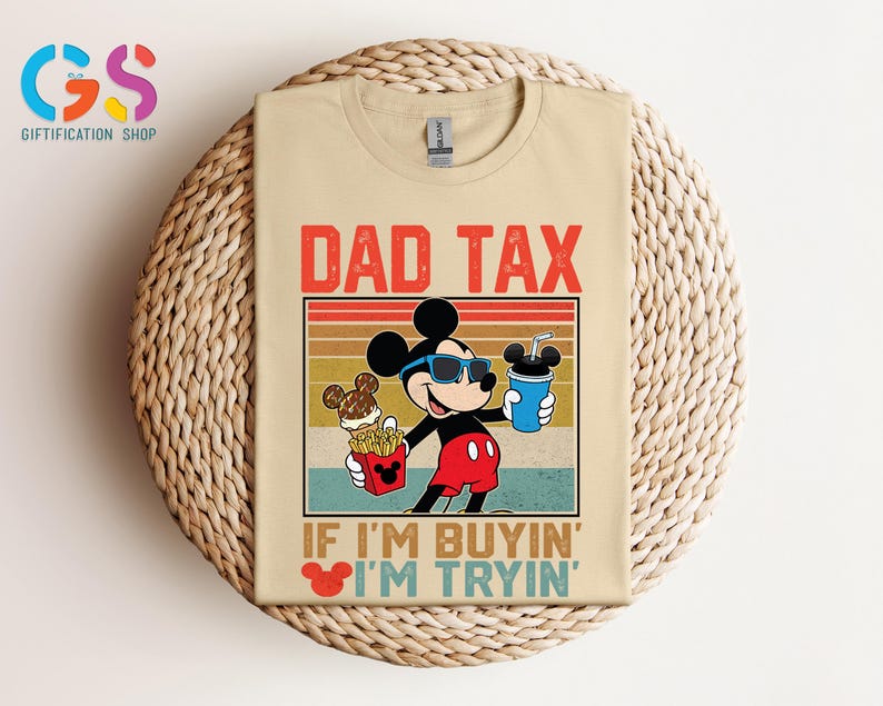 May include: Beige t-shirt featuring a graphic of Mickey Mouse wearing sunglasses and holding a drink and fries. The text "DAD TAX" is above the graphic, and "IF I'M BUYIN' I'M TRYIN'" is below. The shirt is on a woven surface.