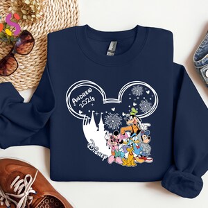 Pode incluir: Camisola azul marinho com um design branco da silhueta do Mickey Mouse, o texto "Andrew 2024" e personagens da Disney. O design inclui fogos de artif&iacute;cio e uma silhueta de castelo. A camisola &eacute; combinada com jeans azuis e sapatos castanhos.