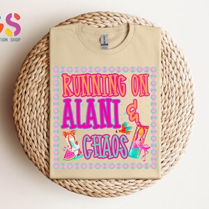 Puede incluir: Una camiseta color beige con las palabras "RUNNING ON ALANI & CHAOS" en rosa, naranja y azul. La camiseta presenta ilustraciones de un loro, un flamenco y una rodaja de sand&iacute;a. La camiseta se muestra sobre una superficie tejida.