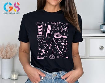 May include: Black t-shirt with a pink graphic design featuring hair styling tools and the words "Hair Stylist". The design includes a barber pole, comb, scissors, and other salon essentials. The shirt is worn by a person.