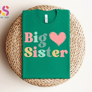 May include: A green t-shirt with the text "Big Sister" in colorful letters and a pink heart.