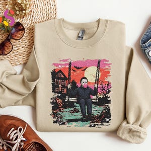 May include: A beige crewneck sweatshirt featuring a graphic print of a character in a white mask sitting on a swing, blowing a pink bubble. The scene includes a house, a large yellow moon, and bats against a pink and orange sunset background.