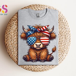 Puede incluir: Camiseta gris claro con un dibujo de una vaca de las Tierras Altas con gafas de sol y una bandana con un dise&ntilde;o de la bandera americana. La vaca tiene pelaje marr&oacute;n y est&aacute; sentada. La camiseta est&aacute; doblada sobre una superficie tejida.