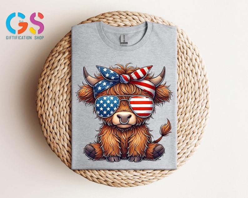 Puede incluir: Camiseta gris claro con un dibujo de una vaca de las Tierras Altas con gafas de sol y una bandana con un dise&ntilde;o de la bandera americana. La vaca tiene pelaje marr&oacute;n y est&aacute; sentada. La camiseta est&aacute; doblada sobre una superficie tejida.
