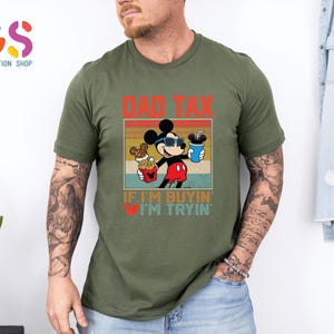 May include: Olive green t-shirt featuring a graphic of Mickey Mouse wearing sunglasses and holding snacks and a drink. The text "DAD TAX" and "IF I'M BUYIN' I'M TRYIN'" are printed above and below the character.