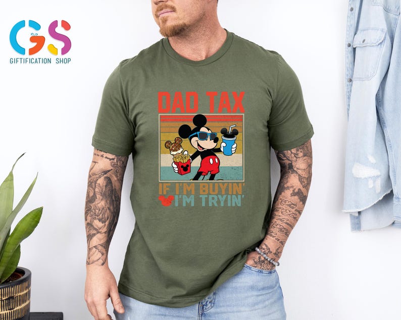 May include: Olive green t-shirt featuring a graphic of Mickey Mouse wearing sunglasses and holding snacks and a drink. The text "DAD TAX" and "IF I'M BUYIN' I'M TRYIN'" are printed above and below the character.