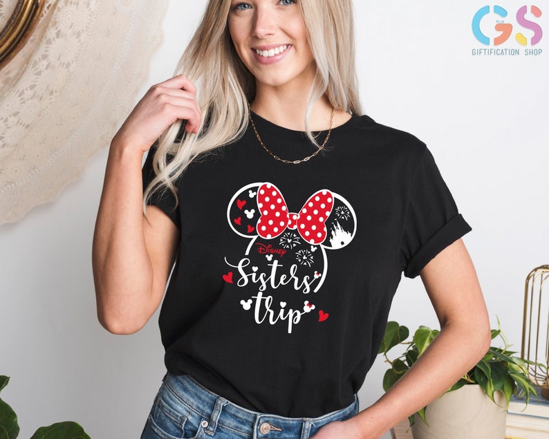 Puede incluir: Camiseta negra con un dise&ntilde;o de orejas de Minnie Mouse blancas y rojas. Las orejas tienen lunares negros y un lazo rojo. El texto "Disney Sisters' Trip" est&aacute; impreso debajo de las orejas.