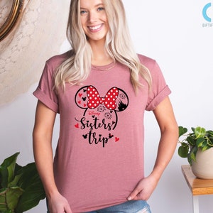 Puede incluir: Una camiseta rosa con un gr&aacute;fico en blanco y negro de orejas de Minnie Mouse con lunares rojos. El gr&aacute;fico tambi&eacute;n incluye el texto "Disney Sisters Trip" en negro.